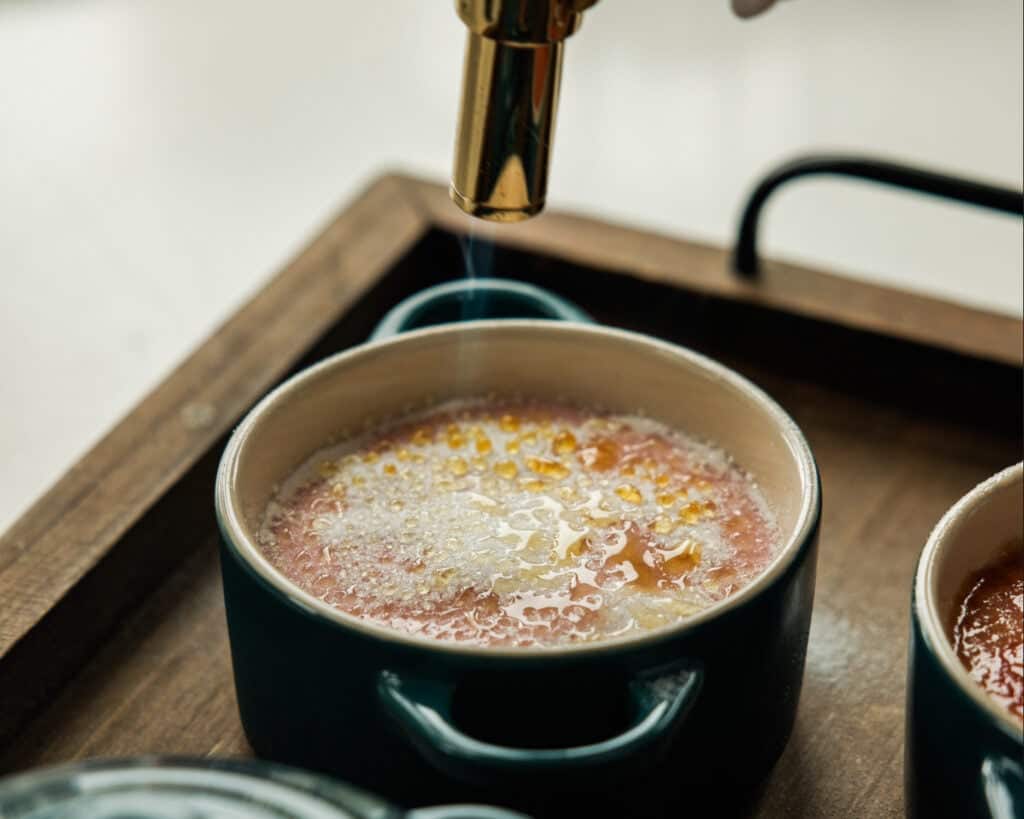 A close-up of a small blue ramekin with Easy Crème Brûlée being caramelized on top using a culinary torch, creating a golden, bubbly crust. The dish sits on a wooden tray.